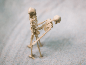 two skeleton figurines over gray background. One skeleton standing while second skeleton bent over in front of first in a standing sex position